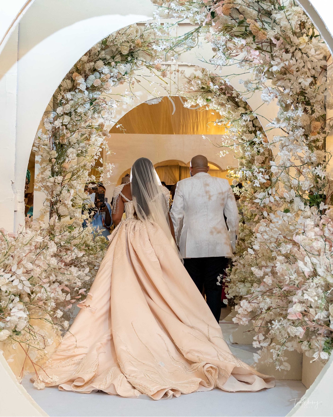 Bride in a blush ballgown with a long train walking through a floral archway with the groom, seen from behind