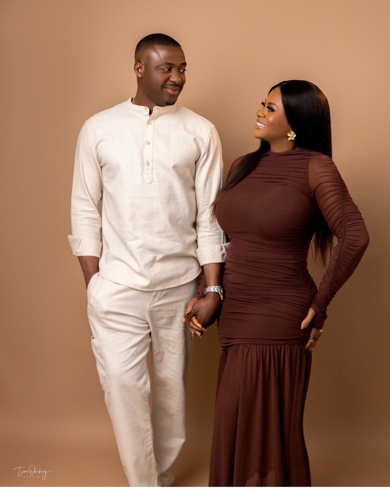 Couple portrait, man in cream traditional wear and woman in brown ruched dress holding hands against a tan backdrop
