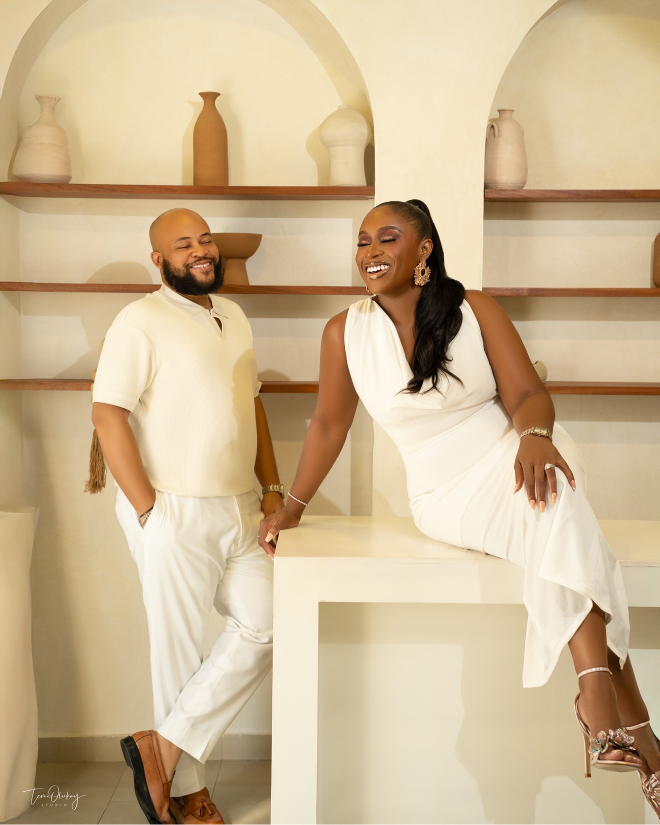 Couple in all-white outfits laughing in bright studio with arched shelving and vases