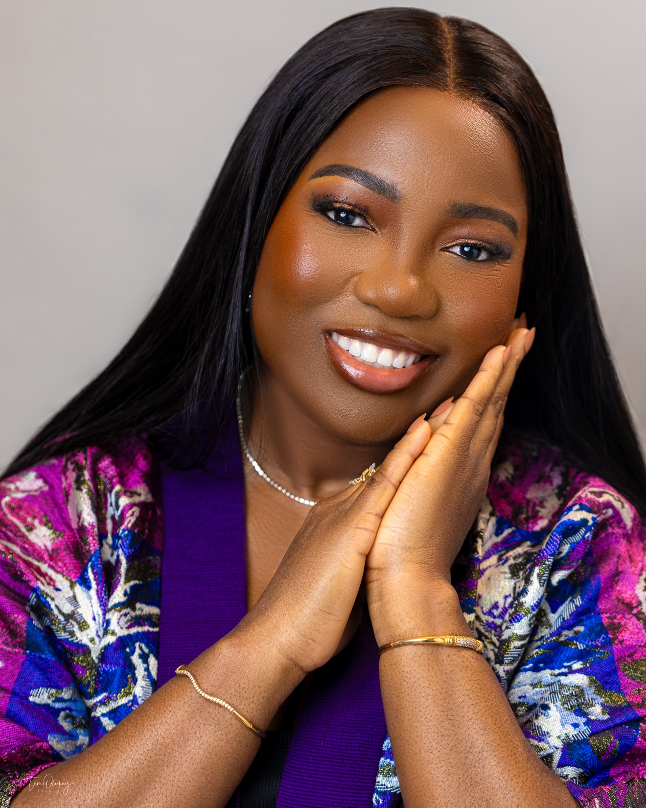 Smiling headshot close-up, colourful purple and pink patterned outfit, hands resting at her face