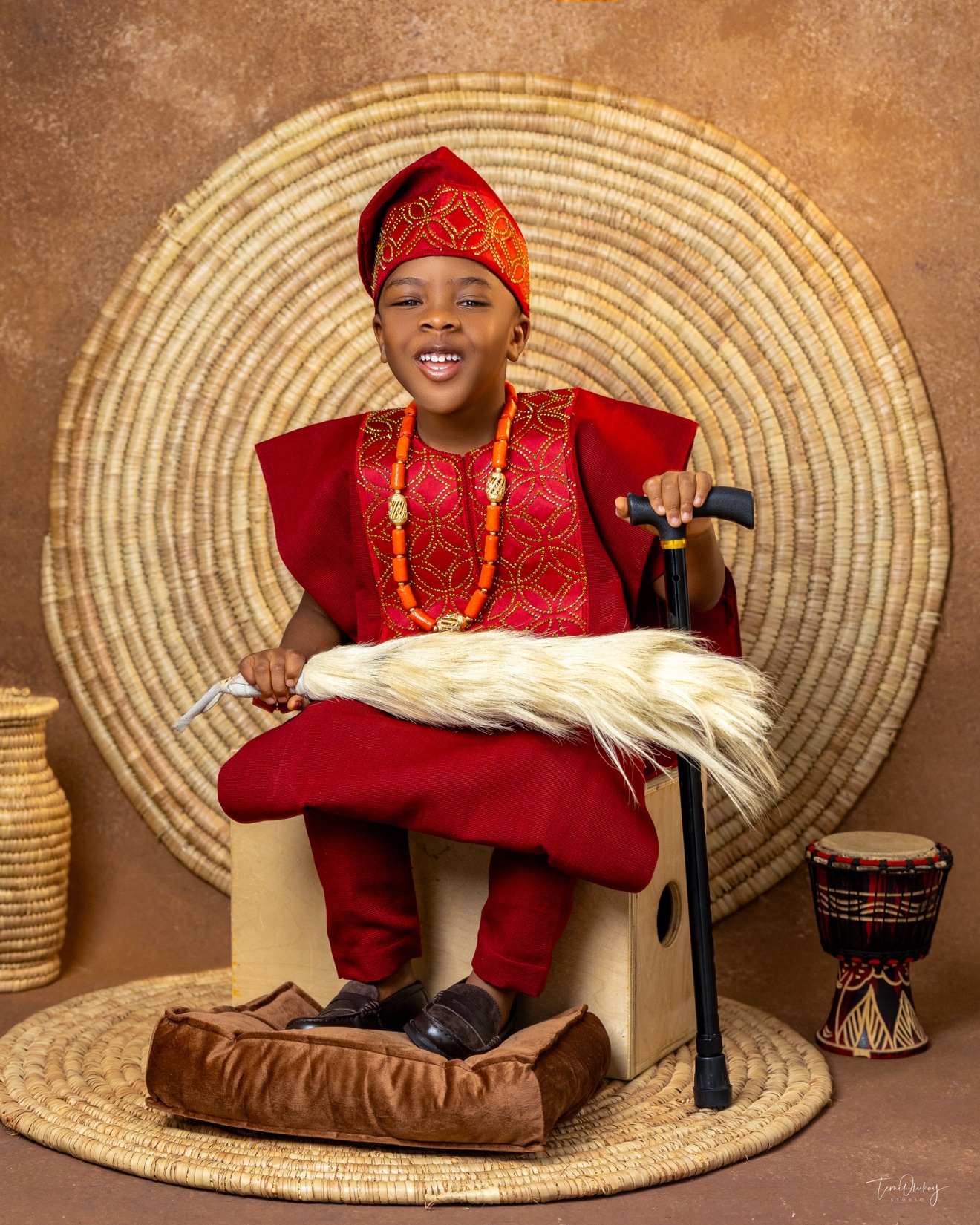 Young boy in red traditional attire with coral beads and cap, woven circular backdrop, holding fly whisk and cane