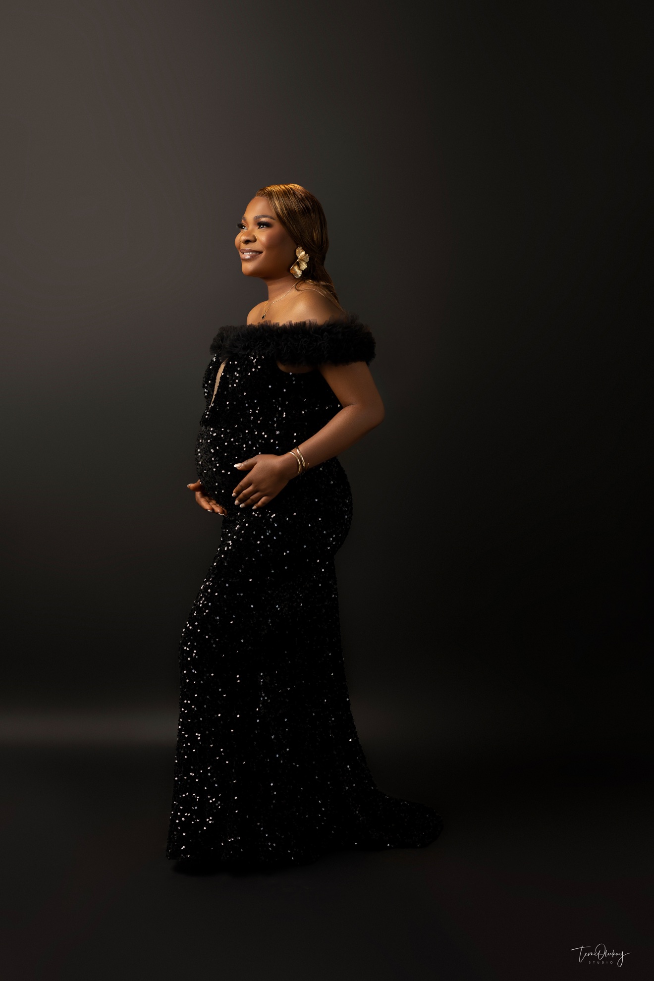 Solo maternity portrait in black sequin gown, profile pose, dark backdrop