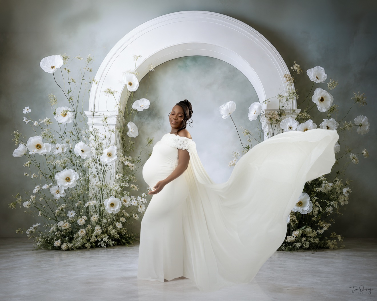 Maternity portrait in flowing white gown with white arch and florals