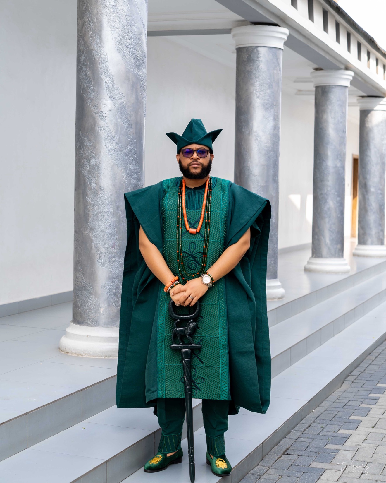 Full-length portrait of a man in a green agbada holding a carved staff, standing by marble columns