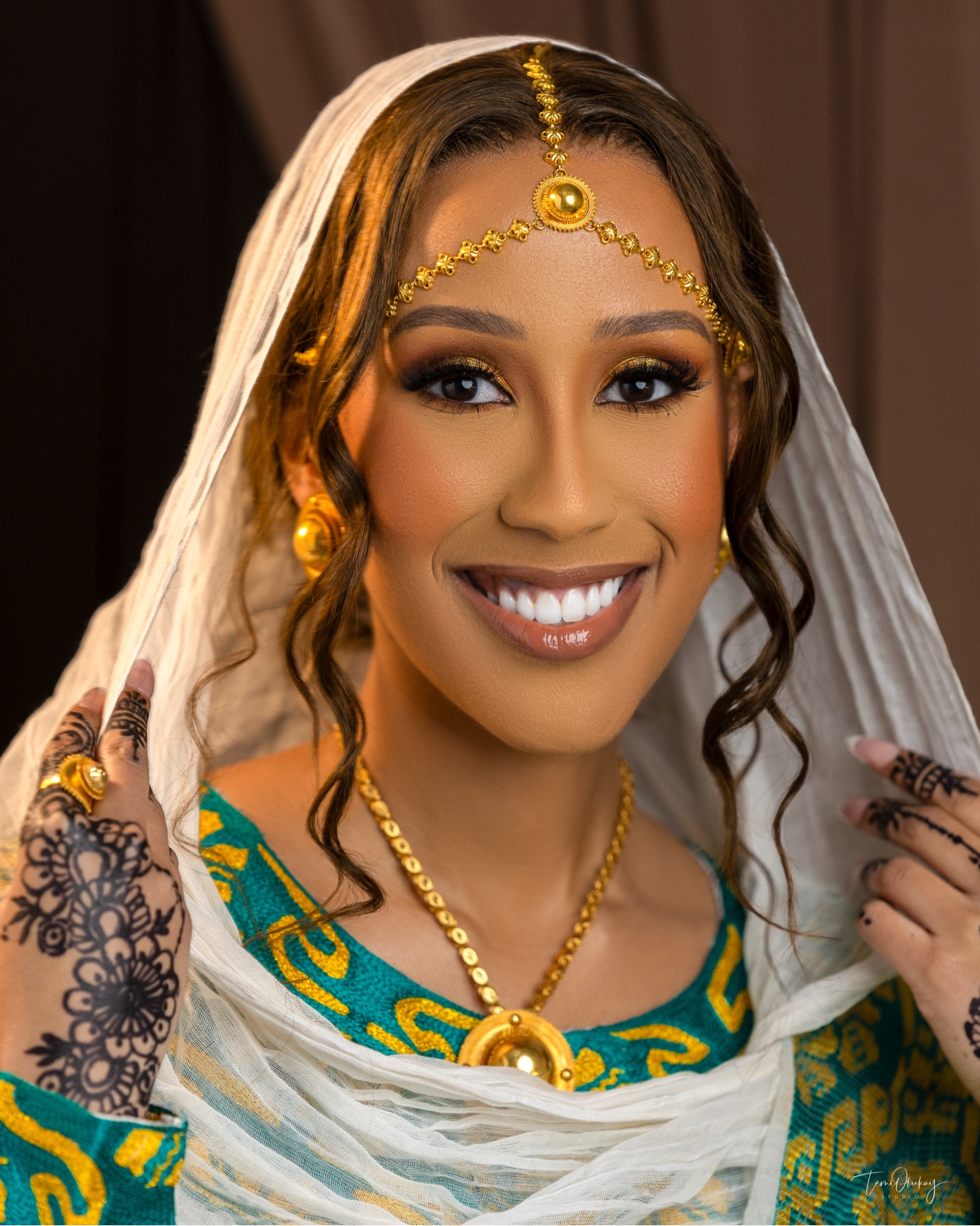 Traditional bride close-up portrait in teal and gold attire with gold jewellery, henna-decorated hands and white veil