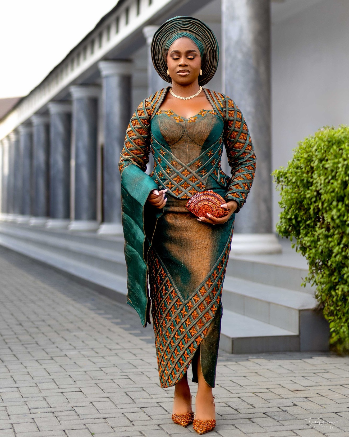 Woman in teal and gold traditional attire with a gele headwrap, standing outdoors by marble columns