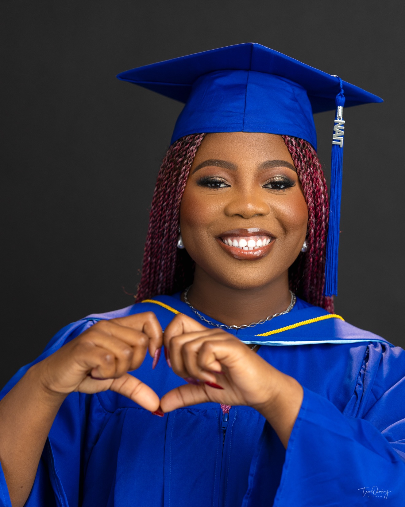 Graduation portrait in blue NAIT cap and gown, making a heart shape with her hands